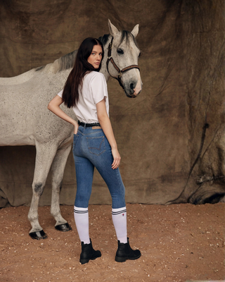 Atmospheric lifestyle shot of a grey horse and model wearing a white t-shirt, OG denim breeches, and white socks.