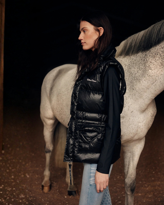 Lifestyle shot of the model from the side with a grey horse, wearing the black puffer vest and black riding top.