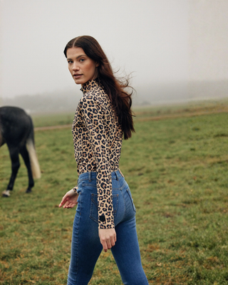 Lifestyle shot of a model walking through a field wearing the Leo riding top and OG denim breeches, looking back.