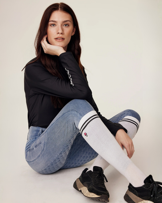 Sitting model wearing a black riding top and light denim breeches, showcasing white riding socks with black stripes and skull detail